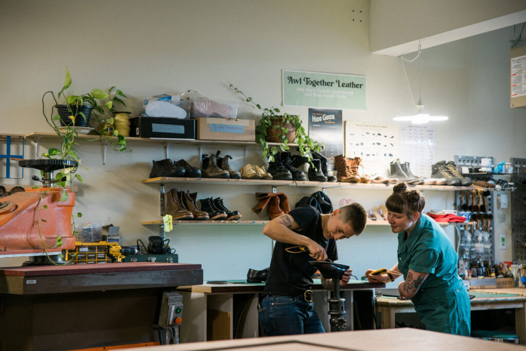 Ariss and Tess in the cobbling area of the shop working on a shoe on a stand. In the background there are plants, machines, tools, and shoes lined up on shelves on the wall.